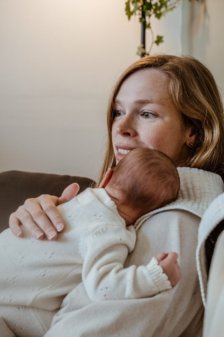 A mother cradles her sleeping newborn against her shoulder in soft daylight.