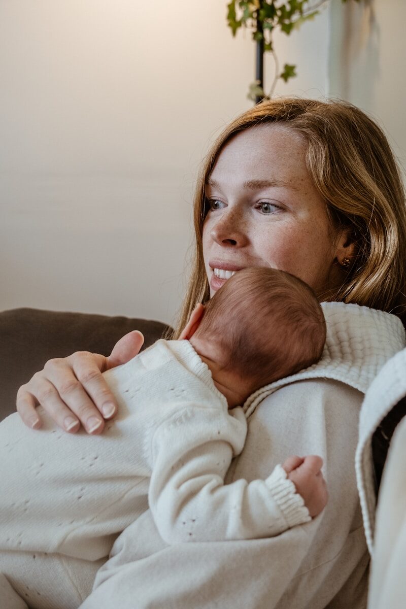 A mother cradles her sleeping newborn against her shoulder in soft daylight.