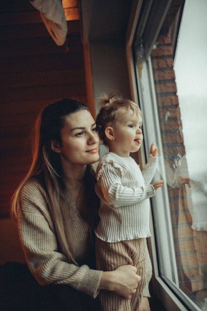 A mother holds her toddler at a bright window, both looking out into the morning light.