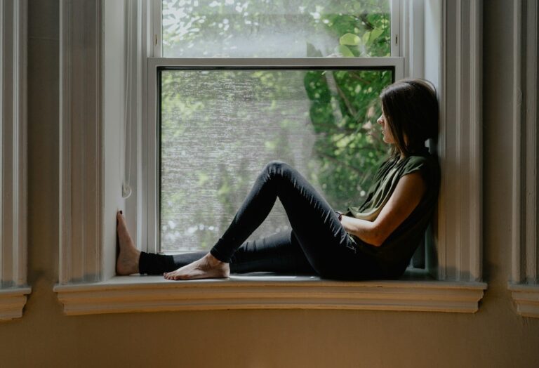 A woman sits on a wide window ledge gazing out at the trees in morning light.