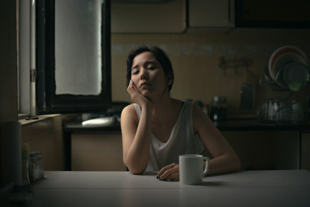 A tired mother sits at her kitchen table late at night, resting her chin on her hand beside a cup of tea.