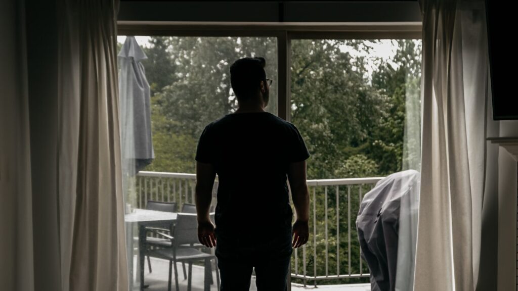 A man stands at an open balcony door of a quiet home, looking out at the trees.