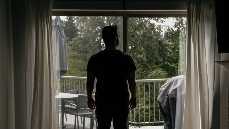 A man stands at an open balcony door of a quiet home, looking out at the trees.
