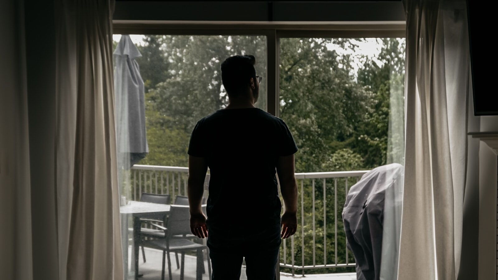 A man stands at an open balcony door of a quiet home, looking out at the trees.