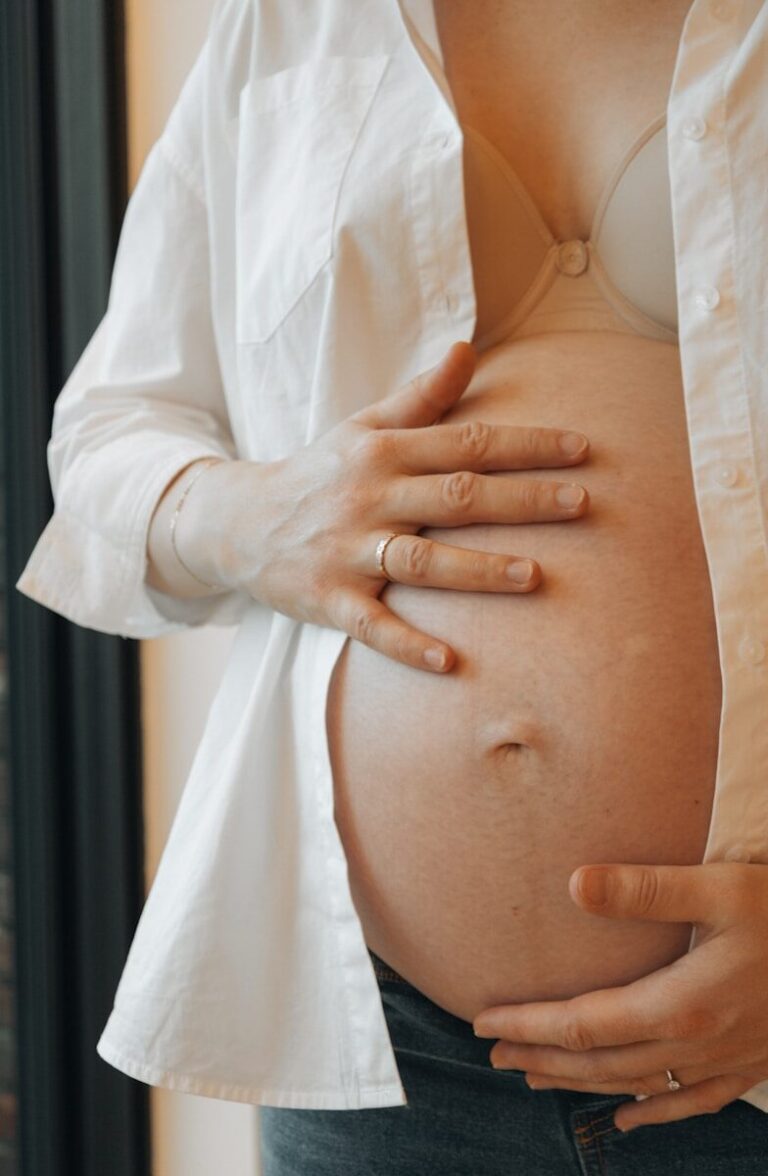 A woman rests her hand gently on her soft bare belly beneath an open white shirt.