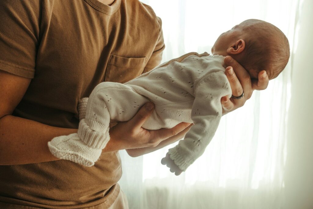 A father cradles his sleeping newborn in his arms beside a sheer-curtained window.