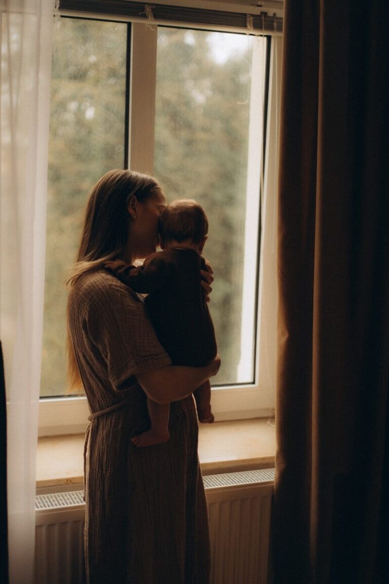 A mother holds her young child close against her chest beside a window in muted light.